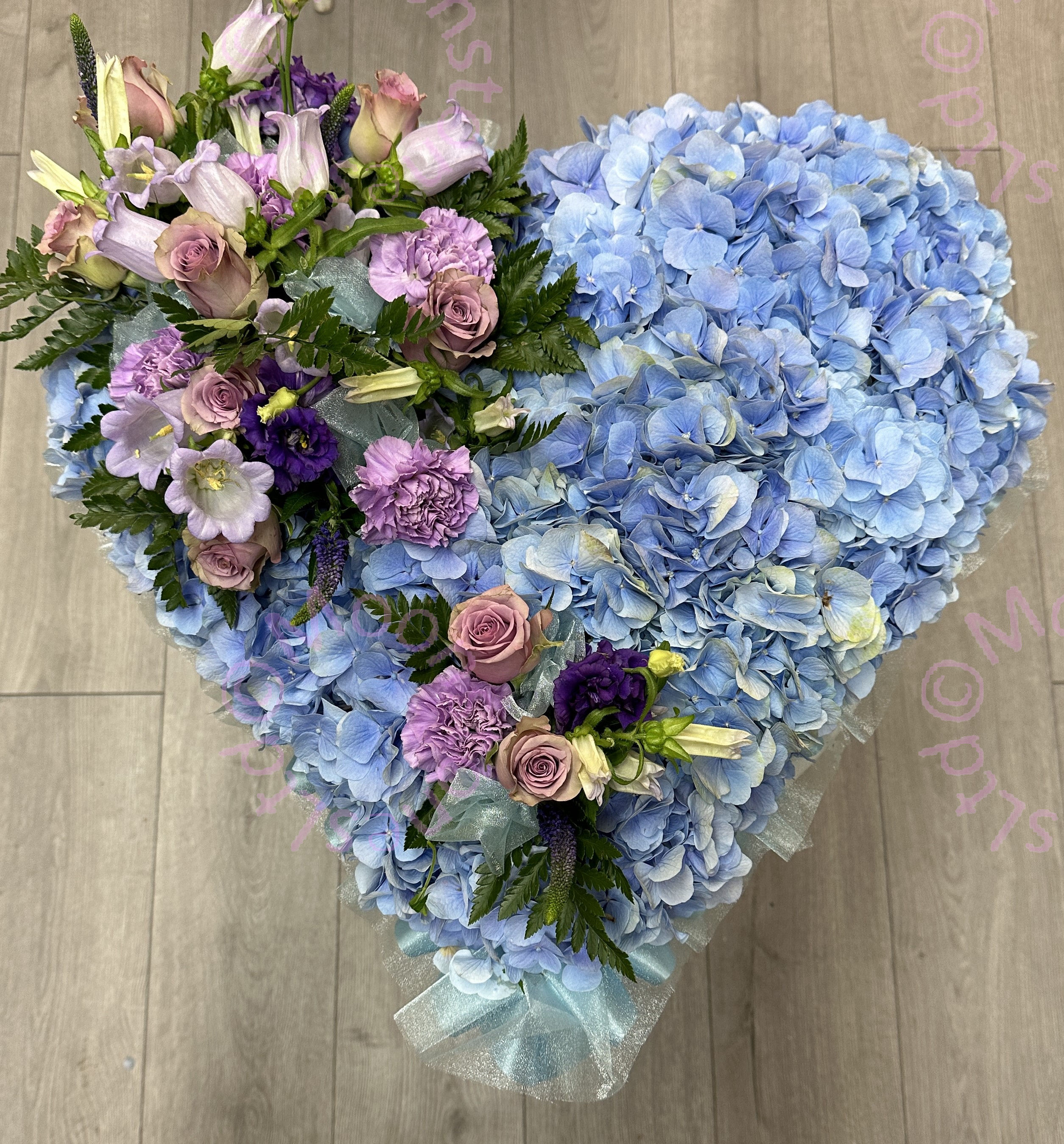 A funeral heart made of pale blue hydrangea with accents of lilac roses. 