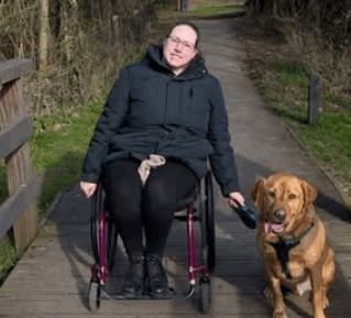 A wheelchair user sitting on a wooden bridge her labrador dog beside her. 
